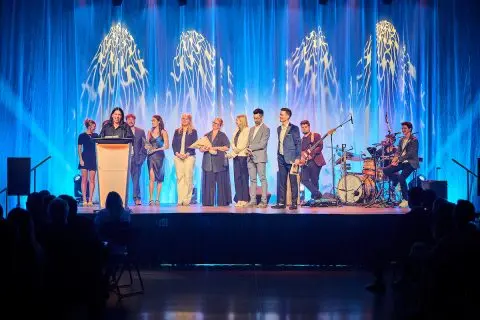 Scène d'un gala de la Chambre de commerce et d'industrie de Sherbrooke (CCIS) où des gens d'affaires sont honorés, avec un orateur au podium, un groupe de musique et un public. Cet événement rassemble les acteurs économiques pour un Sherbrooke innovant et attractif.